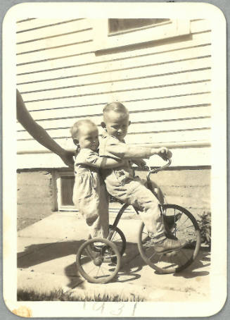 Child riding tricycle with a younger child piggybacking behind him; an adult hand reaches into frame