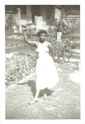 Woman with dark skin tone wearing dress stands in garden with one arm up, holding on to clothesline