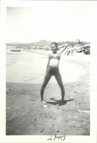 Black-and-white photo of female-presenting person standing in shorts and bikini top on a beach