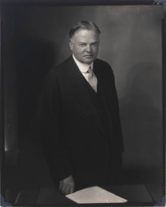 Black-and-white photograph of Herbert Hoover standing before a low table with a small stack of paper