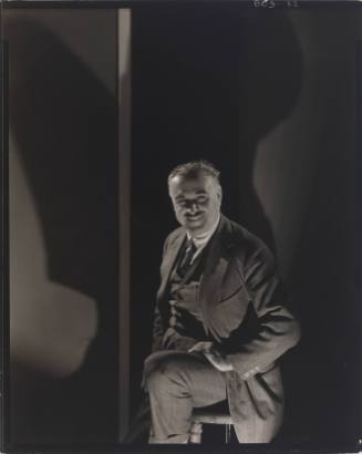 Smiling man in suit with light skin tone and mustache seated on stool with shadow in background