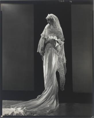 Light-skinned woman standing on pedestal in veil and ornate white gown with train falling to floor