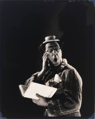 Man with comical, surprised expression wearing small hat, glasses, and plaid coat and holding a book