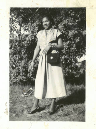 Woman with dark skin tone, wearing dress and heels and holding purse, poses and smiles at camera