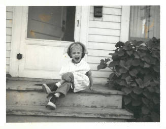 Child wearing heels and clothes that are too big, sitting cross-legged on front porch