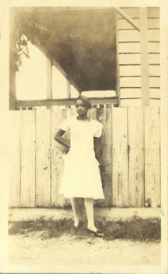Girl with dark skin tone wearing dress poses in front of house with wooden fence, looking at camera
