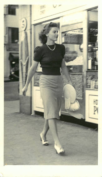 Woman with light skin tone wearing knee-length skirt, sweater, heels, and pearls walks on sidewalk