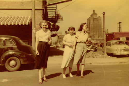 Three women with light skin tone in midi skirts and button-up shirts pose on a car-lined street