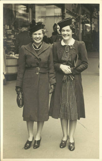 Two women with light skin tone in coats, hats, and heels pose on a crowded city sidewalk