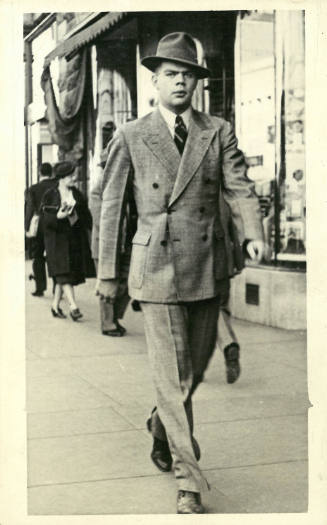 Man in double-breasted suit and hat walking down a busy street in front of storefronts