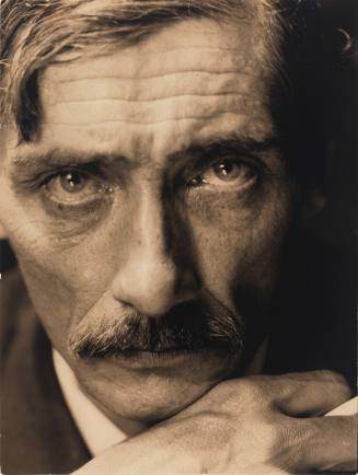 Close-up photo of man with medium light skin tone and mustache looking at camera with hand near face