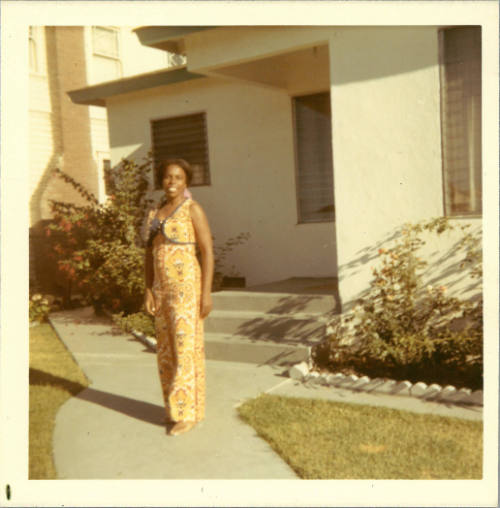 Color photo of smiling woman with dark skin tone wearing yellow dress posing in front of white house