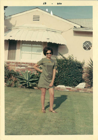 Female-presenting person with medium skin tone in green mini dress smiling in front of a home