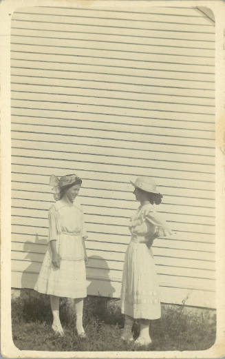Two young women with light skin tone wearing hats and light-colored flowy dresses face each other