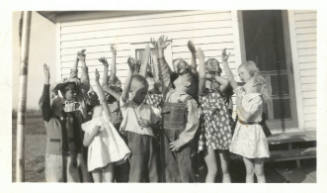 A group of children with light skin tone stand, mostly facing camera, all raising one arm in the air