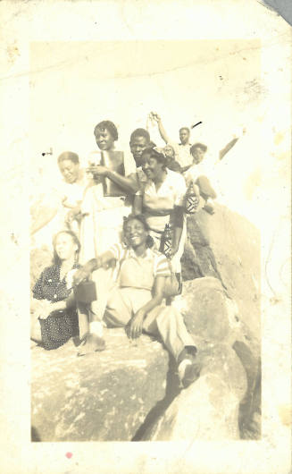 Group of people with dark skin tone, some posing and laughing, sit atop craggy rocks
