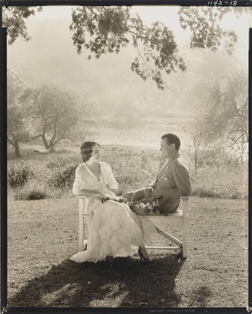 Black-and-white photograph of a woman in a long dress and a man in a suit sharing a bench outdoors
