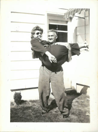 An older man and woman, both with light skin tone and smiling; the man holds the woman in his arms
