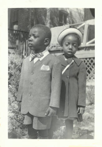 Two young children with dark skin tone, the boy in a blazer and bowtie and a girl in a dress coat