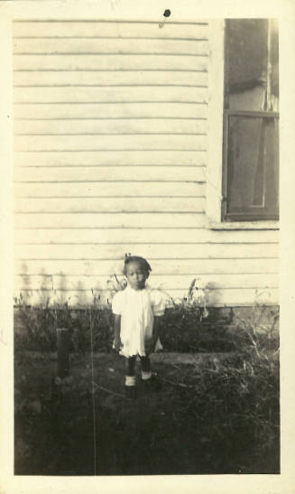 Blurry photograph of young girl with dark skin tone wearing white dress in front of a white house