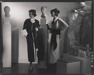 Black-and-white photograph of two women in sleek dresses standing by figural sculptures