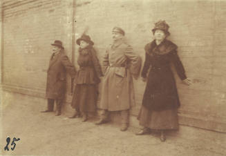 Two men and two women in late 19th or early 20th century clothing stand in line against brick wall