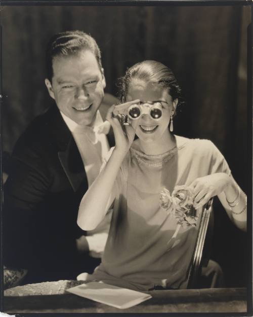 Photograph of smiling, formally dressed light-skinned couple; the woman holds opera binoculars