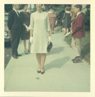Light-skinned woman in shift dress and gloves on crowded sidewalk, possibly in front of a church