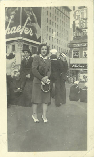 Woman with light skin tone in dress suit standing on city sidewalk in front of men in navy uniforms