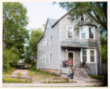 Photograph of a gray, two-story building with driveway at left and two people sitting on the porch