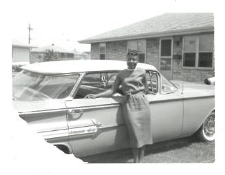 Woman with dark skin tone and bangs in dress standing in front of a car in a driveway