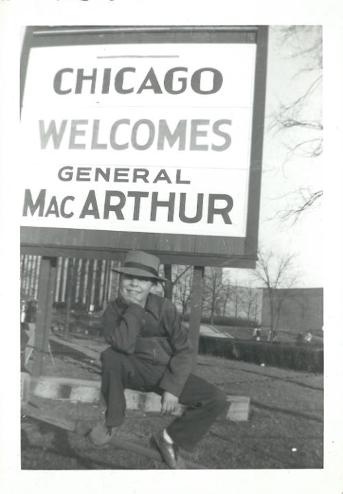 Black-and-white photo of child posing before a sign that reads “CHICAGO WELCOMES GENERAL MacARTHUR”