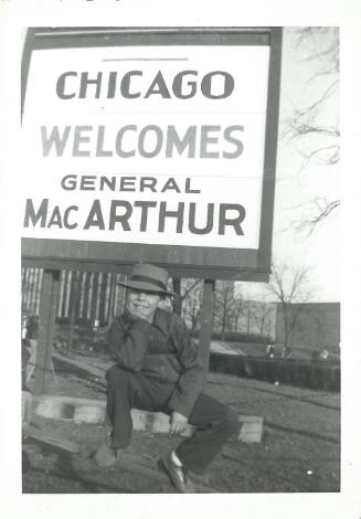 Black-and-white photo of child posing before a sign that reads “CHICAGO WELCOMES GENERAL MacARTHUR”