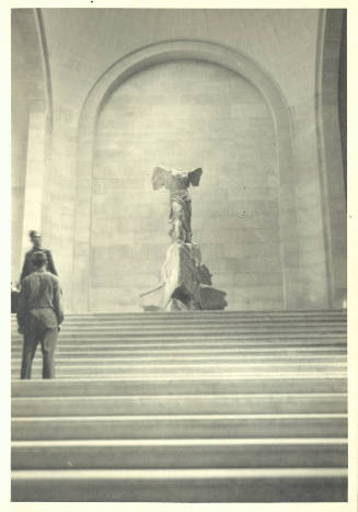 Two people standing on the left side of a staircase and, at top, the Winged Victory of Samothrace