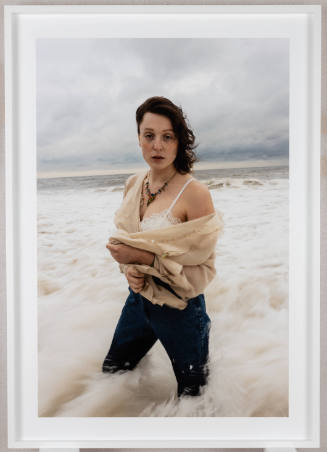 Photograph of fair-skinned person, wearing jeans and an open blouse, standing knee-deep in the ocean
