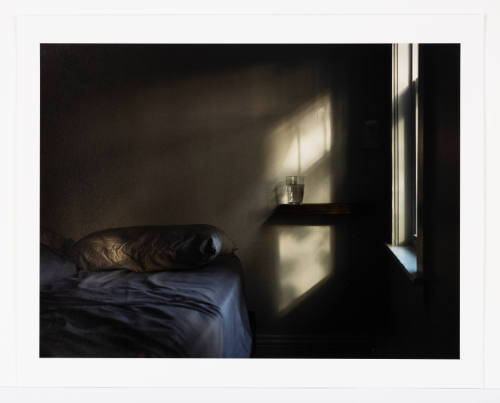 Dim corner of bare-walled room with curtained window, water glass on bedside table & empty bed