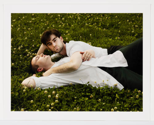 2 light-skinned men in white shirts lie on grass snuggled together, 1 looks up & 1 has eyes closed