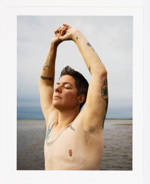 A light-skinned shirtless adult stretches arms up, eyes closed, on beach; tattoos & scars on skin