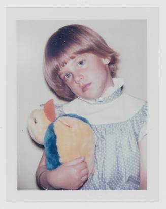 Polaroid of young, blonde child with bowl cut and frilly collar who holds a stuffed yellow duck