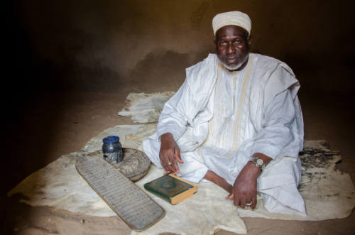 Dark-skinned man in white robe sits on animal hide in dark interior with manuscript and Qur’an board