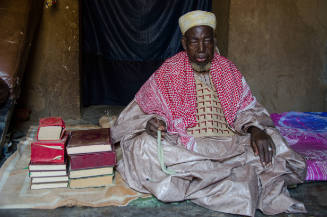 Dark-skinned man in a cap, pink shawl, embroidered robe sits in dark interior next to stacks of book