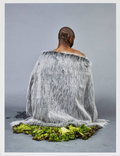 Dark-skinned person with logo shaved onto hair wears faux-fur cape on a bed of lettuce.