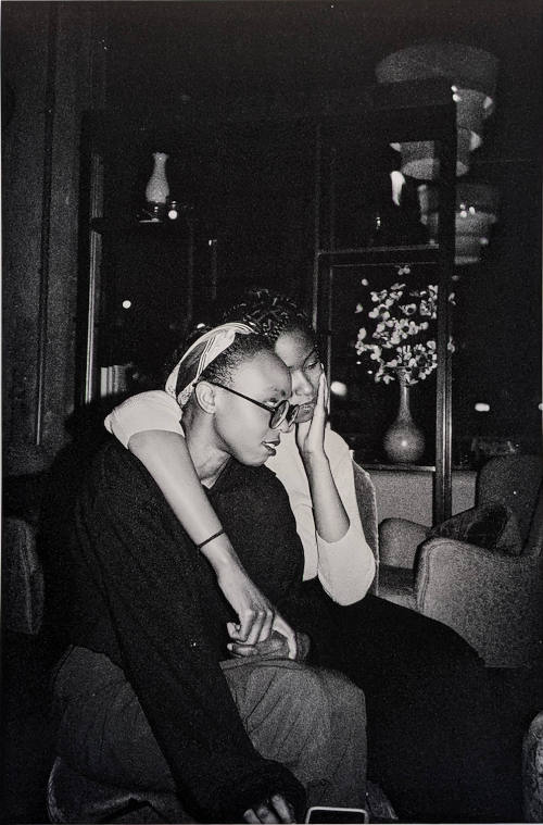 B & W photo of two Black women who embrace & hold hands while sharing a single seat