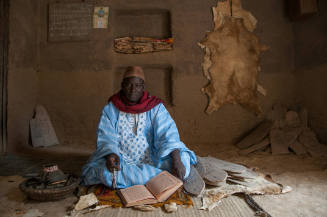 Dark-skinned man in blue embroidered robe sits on mat with manuscript & Qur’an boards stacked behind