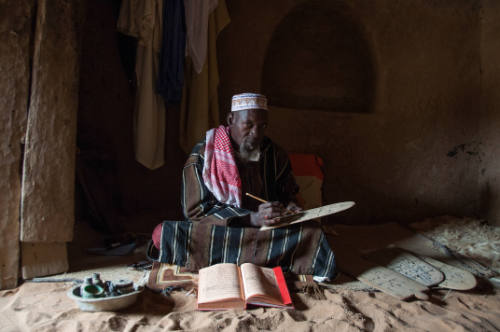 Dark-skinned man in striped robe sits in dark interior and copies from manuscript onto Qur’an board