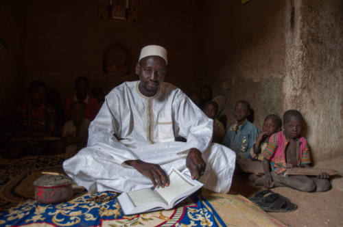 Dark-skinned man in white robe sits on mat with manuscript as boys with Qur’an boards sit behind