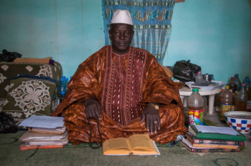 Dark-skinned man in brown embroidered robe sits cross-legged in crowded interior with manuscripts