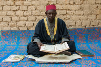 Dark-skinned man in red cap sits on large patterned mat against tan brick wall holding manuscript