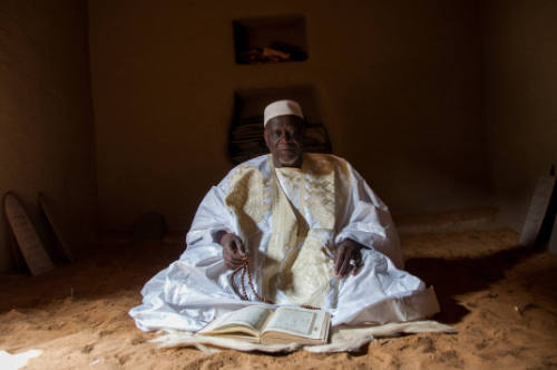 Dark-skinned man in white embroidered robe and cap sits in dark interior with manuscript before him