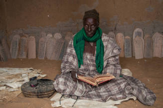 Dark-skinned, robed man sits on animal hide with manuscript; Qur’an boards lined up behind 